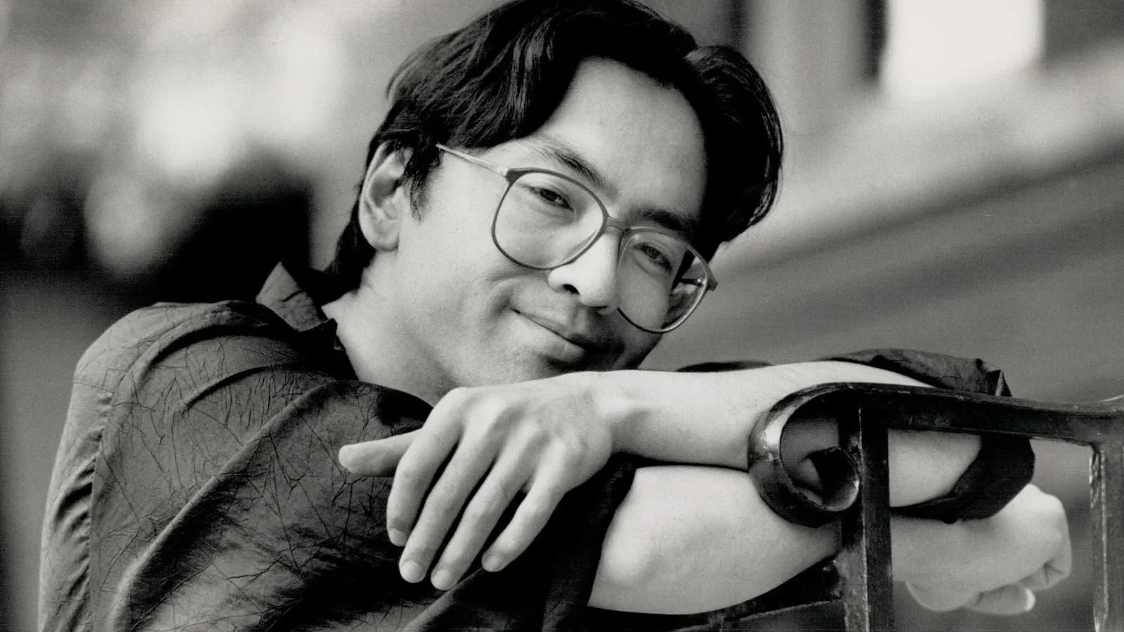 Kazuo Ishiguro photographed early in his career, black and white, leaning on the back of a chair